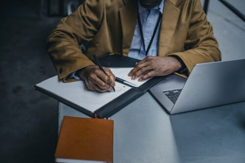 Crop black male executive writing on paper sheet near laptop